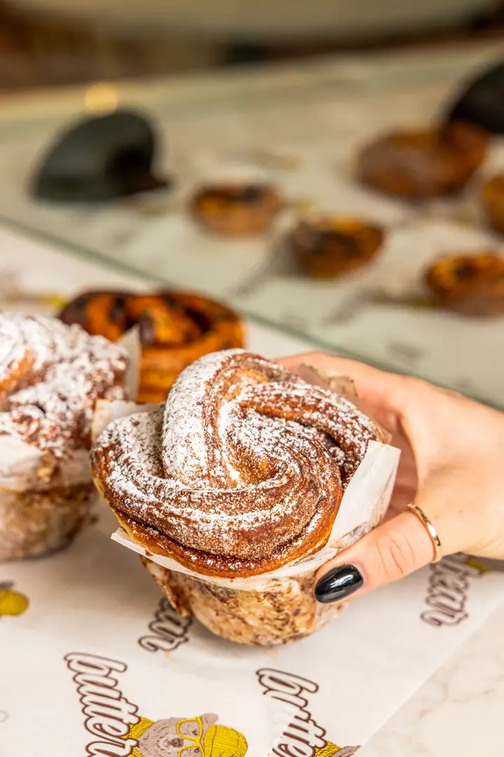 A hand holding a sugar-dusted cruffin above a cabinet of fresh pastries
