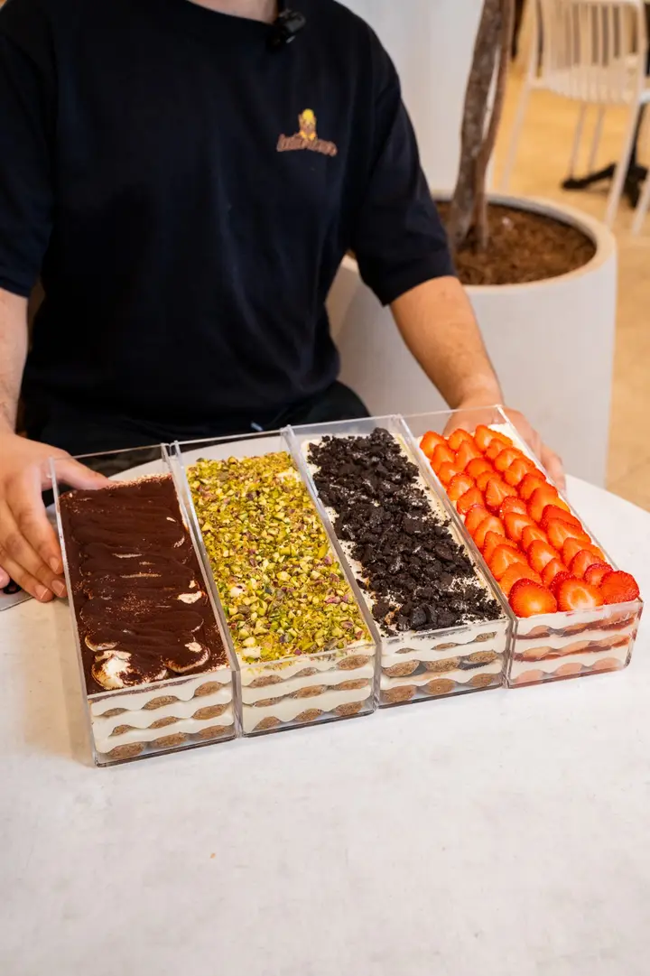 Four long tiramisu trays topped with cocoa, crushed pistachio, cookies and cream, and fresh strawberries